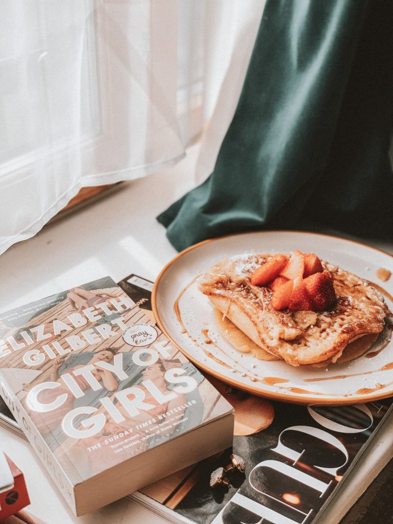 City of Girls book on a window sill with a plate of pancakes. Both positioned on a Vogue magazine. Green curtain